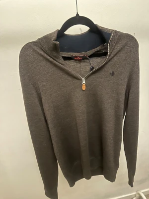 Brun merinoullströja från Morris - Snygg brun långärmad tröja från Morris i 100% merinoull. Tröjan har en liten broderad logga på bröstet och en stilren half zip-dragkedja vid halsen. Perfekt för dig som gillar klassisk och clean stil med en modern touch.