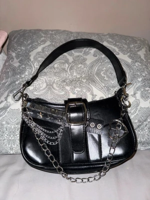 Svart axelväska med kedjedetaljer - Oanvänd cool svart axelväska i skinnimitation med silvriga kedjor, nitar och spänne framtill. Väskan har en bred axelrem och flera snygga metalldetaljer, inklusive hjärtformad ring och stjärnnitar. Perfekt accessoar för en edgy look.