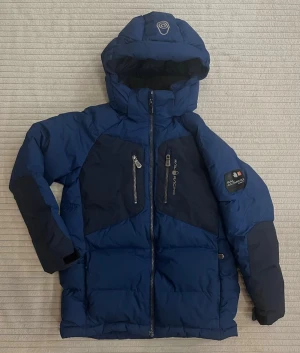 Blå Sail Racing dunjacka med huva, strl 150 - Säljer en blå dunjacka från Sail Racing, modell Antarctica Expedition. Jackan har huva, två stora bröstfickor och två vanliga fickor med dragkedja. Jackan är quiltad och har mörkblå detaljer på axlar och ärmar. Perfekt för kalla vinterdagar! Några små defekter, har fotat dem. 