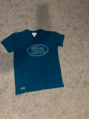Blå Lacoste t-shirt med logga - Snygg blå t-shirt från Lacoste i storlek S. T-shirten har klassisk rund hals, korta ärmar och en stor vit Lacoste-logga med krokodil på bröstet. Tillverkad i mjuk bomull som känns skön mot huden. Perfekt för dig som gillar stilrena märkesplagg.