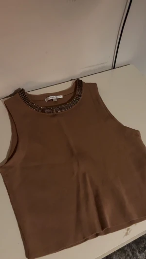 Festlig Brun ärmlös topp med pärlor - Säljer en snygg brun ärmlös topp från Only med pärldetaljer runt halsringningen. Toppen har en rak och croppad passform som ger en cool vibe. Perfekt att styla med jeans eller kjol för en trendig look.