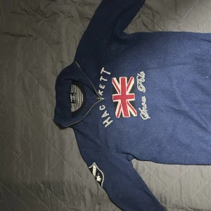 Marinblå merinotröja Hackett London - Snygg marinblå stickad tröja från Hackett London i 100% merinoull. Tröjan har half zip, lång ärm och coolt broderad Union Jack-flagga samt texten 'Hackett' och 'Aston Polo' på bröstet. Patch på ena ärmen ger extra stilpoäng.
