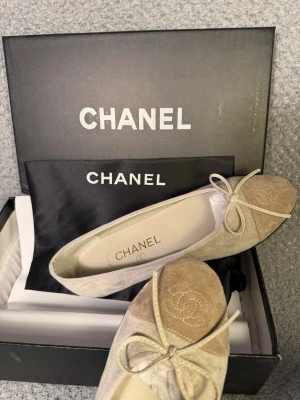 Beige ballerinaskor från Chanel - Snygga ballerinaskor från Chanel i beige mocka med rund tå och en elegant rosett framtill. Tåpartiet är i en mörkare beige ton med broderad Chanel-logga. Skorna har platt sula och klassisk design, perfekta för dig som gillar lyxiga detaljer.