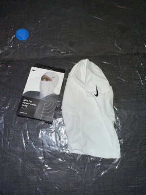Vit Nike Pro Hyperwarm Hood - Nike Pro Hyperwarm Hood i vit med svart Nike-logga. Snygg och funktionell balaclava som täcker hela huvudet och halsen, perfekt för kalla dagar. Tillverkad i mjukt och stretchigt syntetmaterial som andas och håller värmen.