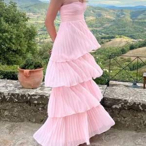 Balklänning - Superfin ljusrosa långklänning med plisserade volanger i flera lager. Axelbandslös modell med hjärtformad urringning och dragkedja bak. Klänningen har en romantisk och flowy känsla, perfekt för speciella tillfällen. Materialet är lätt och ger ett drömmigt intryck.
