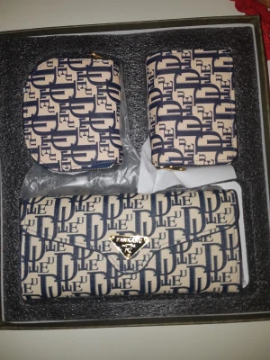 Beige och blå plånboksset från Fankanu - Snyggt set med tre plånböcker från Fankanu i beige och mörkblått med grafiskt mönster. Setet innehåller en större kuvertväska med guldfärgad logga, en mindre plånbok och en myntbörs, alla med dragkedja och matchande design. Perfekt för att hålla koll på kort, kontanter och småsaker.