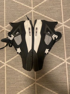 Svarta Nike Air Jordan 4 sneakers - Säljer ett par svarta Nike Air Jordan 4 sneakers med vita och grå detaljer. Skorna har meshpaneler på sidorna, snörning och en synlig Air-sula. Ikonisk Jumpman-logga på plösen och 'Flight'-text. Materialet är en mix av skinn och syntet, med platt sula och rund tå.