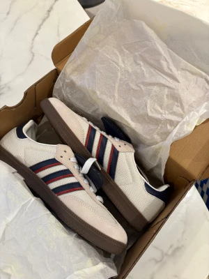 Adidas Samba OG crystal white - Säljer ett par Adidas Samba OG med vit mesh och beige mockadetaljer, klassiska marinblå och röda stripes på sidorna samt brun gummisula. Skorna har snörning och rund tå, perfekt för dig som gillar retrovibbar och streetstyle.