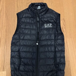 Svart dunväst från EA7 Emporio Armani - Snygg svart dunväst från EA7 Emporio Armani med quiltad design och hög krage. Västen har dragkedja framtill och två sidofickor. Logotypen är broderad på bröstet för en exklusiv touch. Perfekt att slänga över en hoodie för en cool look.