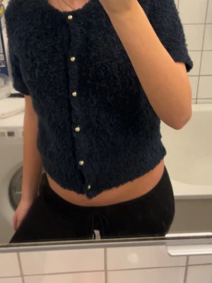 Mörkblå fluffig kofta från Gina Tricot - Supermysig mörkblå kofta från Gina Tricot med korta ärmar och croppad passform. Koftan är fluffig och har guldiga knappar framtill som ger en snygg detalj. Perfekt för dig som gillar mjuka och trendiga plagg.