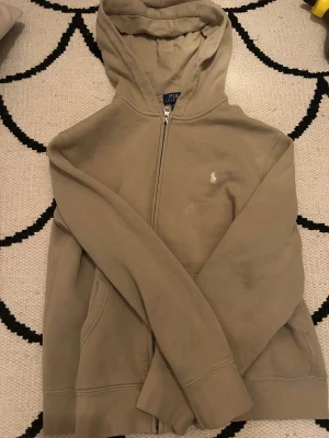Ralph lauren zip hoodie - En beige hoodie från ralph lauren med zip.Den har ett vit Ralph lauren märke på vänster sida.Den är i väldigt bra skick har en väldigt skön och varm insida 