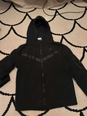 Nike tech fleece - En svart Nike fleece med två streck som går diagonalt från dragkedjan.Den har en smart ficka på sin vänster arm med blixtlås.Den har tvättats i fel grad därför har Nike märket gått upp längst nere lite grann.