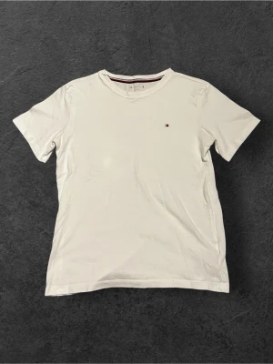 Vit t-shirt från Tommy Hilfiger - Vit t-shirt från Tommy Hilfiger med klassisk rund hals och diskret logga broderad på bröstet. Halsringningen har en snygg detalj med röd, vit och marinblå rand. Tillverkad i mjuk bomull för skön känsla hela dagen.