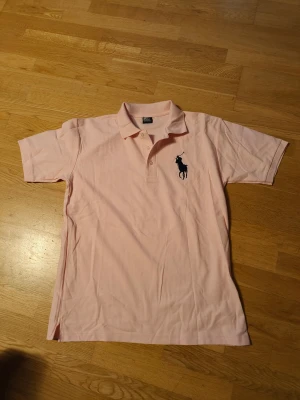 Rosa polotopp från Ralph Lauren - Säljer en ljusrosa polotopp från Ralph Lauren med klassisk krage och knappar framtill. T-shirten har korta ärmar, ett stort mörkblått Polo-logotryck på bröstet och en siffra på ena ärmen. Materialet är mjuk bomull som känns skön mot huden.