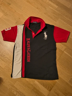 Ralph Lauren svart och röd polotopp - Säljer en snygg polotopp från Ralph Lauren med svart och röd färgblock, vit detalj på sidan och stor logga på bröstet. Toppens krage är röd med  text, och ärmarna är korta med vita siffror på ena sidan. Perfekt för dig som gillar sportig stil.