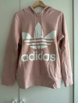 Rosa Adidas hoodie med logga - Säljer en rosa hoodie från Adidas med det klassiska trefoil-loggan i vitt framtill. Tröjan har huva med snörning och långa ärmar. Perfekt för både vardag och chill dagar
