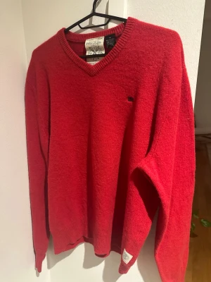 Röd ulltröja Polo Jeans Ralph Lauren - Riktigt snygg röd tröja från Polo Jeans Ralph Lauren i 100% lammull. Klassisk v-ringad modell med ribbade muddar och diskret logga på bröstet samt patch på ärmen. Perfekt för dig som vill ha en stilren och tidlös look.