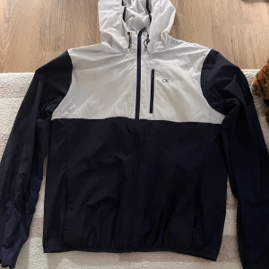 Calvin Klein svartvit vindjacka - Snygg vindjacka från Calvin Klein i svart och vitt med huva och dragkedja framtill. Jackan har en bröstficka med dragkedja, justerbar dragsko i huvan och diskret CK-logga på bröstet. Perfekt för blåsiga dagar och stilren streetlook.