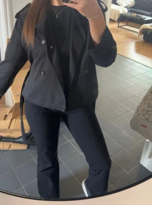 Svart kort trenchcoat Stradivarius - Snygg svart kort trenchcoat från Stradivarius med klassisk krage, knappar och bälte i midjan. Jackan har axelklaffar och långa ärmar, perfekt för dig som vill ha en stilren look. Materialet är mjukt och lätt, vilket gör den skön att bära under vår och höst.