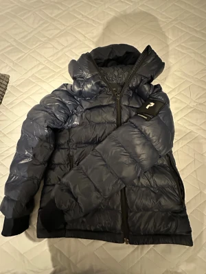 Mörkblå pufferjacka från 1 More - Säljer en mörkblå pufferjacka från 1 More med huva och svarta muddar vid ärmslut. Jackan har en snygg glansig yta, dragkedja framtill och en patch med logga på ena ärmen. Perfekt för kalla dagar och riktigt skön att bära. Pris kan diskuteras, har bara en liten mini skada som inte syns på ryggen.