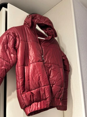 Vinröd pufferjacka med huva - Snygg vinröd pufferjacka med glansig finish och quiltad design. Jackan har en stor huva, halv dragkedja med cool ringdetalj och breda muddar nertill. Perfekt för kalla dagar när du vill hålla stilen och värmen. Storlek S men passar lika bra XS🥰helt oanvänd, endast provad och köptes för 800 kr.