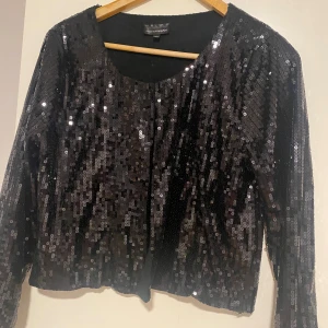 Svart paljettkofta från KappAhl - Glittrig svart kofta från KappAhl med paljetter över hela plagget. Rund halsringning och långärmad modell som ger en snygg och festlig look. Perfekt för dig som vill sticka ut med lite extra bling. Snygg att matcha med svarta jeans eller kjol.