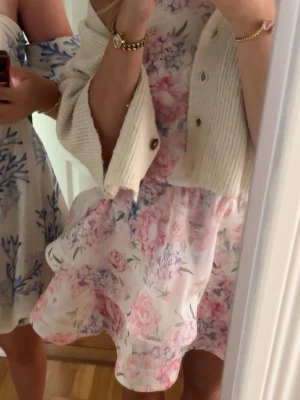 Blommig kortklänning i pastellfärger - Supersöt kortklänning med volang och blommigt mönster i ljusa toner av rosa, lila och vitt. Klänningen har en rund hals och är ärmlös, perfekt för sommarens alla tillfällen. Materialet är lätt och luftigt, vilket gör den extra skön att bära.