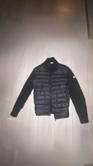 Svart Moncler pufferjacka med stickade ärmar - Svart pufferjacka från Moncler med quiltad front och stickade ärmar. Jackan har hög krage, dragkedja och Moncler-logga på vänster ärm. Materialet är en mix av dun och stickad polyester, perfekt för dig som gillar stilren och sportig vibe.