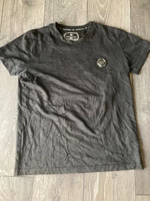 Svart Philipp Plein t-shirt med emblem - Svart t-shirt från Philipp Plein, Platinum Cut, med coolt metall-emblem på bröstet. T-shirten har korta ärmar och ett diskret prickigt mönster i tyget. Materialet är mjuk bomull och passformen är slim. Perfekt för dig som gillar exklusiva streetwear-vibbar.