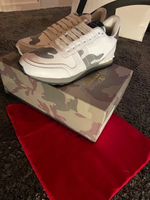 Valentino Rockrunner  - Snygga Valentino Rockrunner sneakers i vitt och grått med camouflagemönster. Skorna har rund tå, platt sula och coola nitar på hälen. Tillverkade i skinn och textil med vita snören och detaljerad design. Perfekt för dig som vill sticka ut med exklusiva sneakers. Pris kan diskonteras 