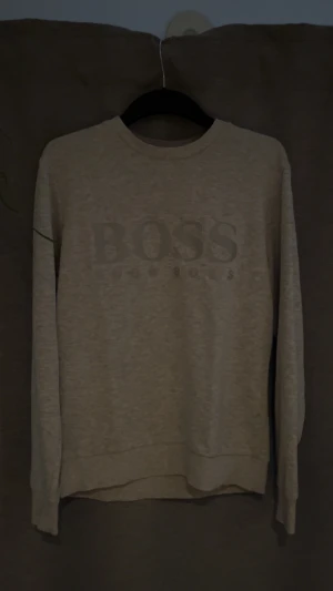 Grå sweatshirt från Hugo Boss - Snygg grå sweatshirt från Hugo Boss med stort BOSS-tryck på bröstet. Märket är i reflex. Tröjan har rund hals, långa ärmar och en klassisk passform. Perfekt för dig som gillar stilrena och enkla designer med en tydlig logga framtill.