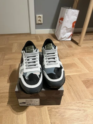 Valentino Garavani Rockrunner sneakers - Säljer ett par Valentino Garavani Rockrunner sneakers i svart, grått och vitt med snyggt kamouflagemönster. Skorna har snörning, platt sula och detaljer i mocka och skinn. Ikonisk logga på plösen och innersulan. Perfekt för dig som gillar exklusiva streetwear-vibes.