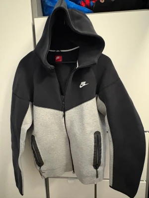 Nike hoodiejacka svart och grå - Nike hoodiejacka i svart och ljusgrå färgblock med dragkedja framtill och två fickor med svarta detaljer. Jackan har stor huva och långärmade ärmar, tillverkad i mjuk syntet och bomullsmix. Snyggt Nike-logga broderad på bröstet.