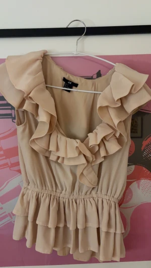 ljusrosa volangblus från H&M - Säljer en ljusrosa blus från H&M med stora volanger runt halsen och axlarna💓. Blusen är ärmlös och har ett elastiskt midjeparti med dubbla volanglager nedtill. Perfekt för dig som gillar romantisk och feminin stil. Ser mer beige ut på bilden alldeles mer ljusrosa i ljuset, kom privat för mer noga bilder 💓Inte några skavanker då den är använd få gånger.