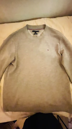 Beige stickad tröja Tommy Hilfiger - Snygg beige stickad tröja från Tommy Hilfiger i storlek L. Tröjan har ett diskret Hilfiger-logo broderat på bröstet och klassisk rund hals. Materialet är mjukt och strukturerat, perfekt för dig som gillar stilrena och enkla plagg. Passar med de mesta 