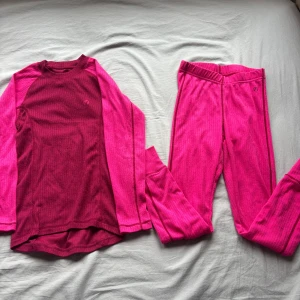 Rosa underställströja och byxor från North Bend - Underställströja och byxor från North Bend i en snygg kombination av mörkrosa och ceriserosa. Tröjan är ribbad och har långa ärmar, perfekt för kyliga dagar. Materialet är mjukt och stretchigt, vilket gör den skön att ha på sig under vinterns aktiviteter.