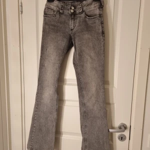 Grå bootcut jeans med knappar - Snygga grå jeans med bootcut passform och cool tvättad look. Framtill finns dubbla knappar och klassiska fickor, baktill har jeansen fickor med lock och knapp. Perfekta för dig som gillar en retro vibe och vill sticka ut med din stil.