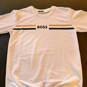 Vit tröja från BOSS - Vit tröja från BOSS med svarta och beige ränder över bröstet och BOSS-logga i svart. Tröjan har rund halsringning och ribbade muddar vid ärmslut och nederkant. Perfekt för en clean och stilren look.