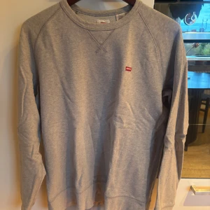 Grå sweatshirt från Levi's i bomull - Säljer en stilren grå sweatshirt från Levi's i mjuk bomull. Tröjan har rund halsringning, långa ärmar och en liten röd Levi's-logga på bröstet. Perfekt för en chill och avslappnad look.