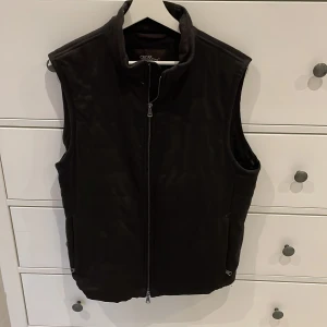 Svart väst från Oscar Jacobson - Snygg svart väst från Oscar Jacobson med hög krage och hel dragkedja framtill. Västen har en stilren design med diskreta fickor på sidorna och lätt quiltade detaljer på ryggen. Perfekt att slänga över en hoodie eller tröja när det är kyligt ute.