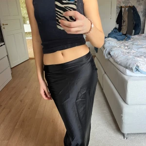 Svart långkjol i satin-look - Säljer en svart långkjol med snygg satin-finish. Kjolen har en slät och glansig yta som ger en stilren vibe. Den är rak i modellen och sitter snyggt i midjan. Perfekt att styla med crop tops eller stickade tröjor.