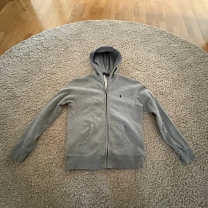 Grå hoodie från Polo Ralph Lauren - Säljer en grå hoodie från Polo Ralph Lauren i storlek L (barnstorlek) motsvarar ungefär XS i vuxenstorlek. Tröjan har huva, dragkedja framtill och två fickor. Klassisk broderad logga på bröstet. Skicket är bra, inga fläckar och sparsamt använd.