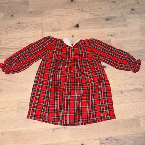 Röd rutig klänning med vit krage - Supergullig röd rutig klänning med lång ärm och vit krage. Klänningen har klassiskt rutmönster i rött, svart och vitt, samt knäppning bak. Perfekt för dig som gillar retrovibbar och vill sticka ut lite extra.Röd, rutig klänning med lång ärm och vit krage. Klänningen har rynkdetaljer vid bröstet och ärmsluten. 🎄 