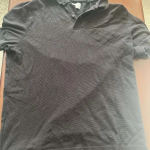 Svart henley från Primark, medium - Svart henley från Primark i regular fit. Skjortan har korta ärmar, klassisk krage och knappar vid halsen. Tillverkad i bomull med diskret textur i tyget. Perfekt för en clean och enkel stil.