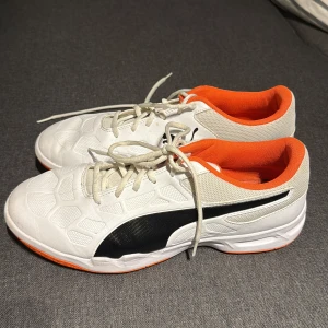 Vita och orange sneakers från Puma - Säljer ett par fräscha sneakers från Puma i vitt med svart logga och detaljer samt orange insida och sula. Skorna har snörning och är tillverkade i syntetmaterial och mesh för bra andningsförmåga. Perfekta för dig som gillar sportig stil.