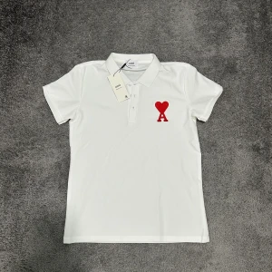 Vit piké från Ami med rött hjärta - Säljer en stilren vit pikétröja från Ami med klassisk krage och knäppning framtill. Tröjan är i mjuk bomull och har korta ärmar. Perfekt för dig som gillar clean och snygg design. Helt ny har inte använt den. Priset kan diskuteras.