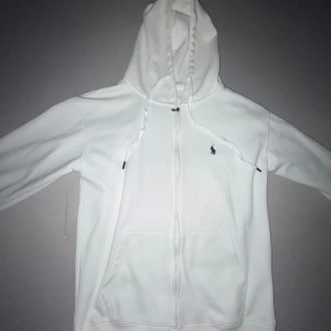 Vit hoodie från Polo Ralph Lauren - Snygg vit hoodie från Polo Ralph Lauren med dragkedja och klassisk logga på bröstet. Tröjan har huva med snörning, känguruficka och är tillverkad i 100% bomull. Perfekt för en clean och stilren look.