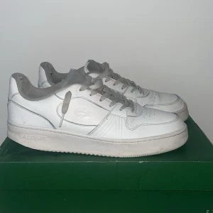 Vita sneakers från Lacoste - Säljer ett par fräscha vita Lacoste sneakers med grå detaljer och snörning. Skorna har perforerade partier på sidorna och tån för extra ventilation, samt en platt sula och rund tå. Klassisk Lacoste-logga på sidan och mjukt foder för skön komfort.