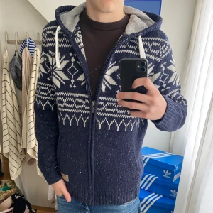 Mörkblå stickad hoodie från Reserved - Säljer en mörkblå stickad hoodie från Reserved i storlek XL men sitter som en L/M .Tröjan har vitt nordiskt mönster, dragkedja framtill, huva med snörning och ribbade muddar. Perfekt för kalla dagar och riktigt snygg till jeans.