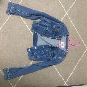 Blå jeansjacka från NAKD  - Säljer en klassisk blå jeansjacka från NAKD i storlek 32. Jackan har silverfärgade knappar, två bröstfickor med lock och långa ärmar. Perfekt att slänga över en hoodie eller t-shirt för en chill look.